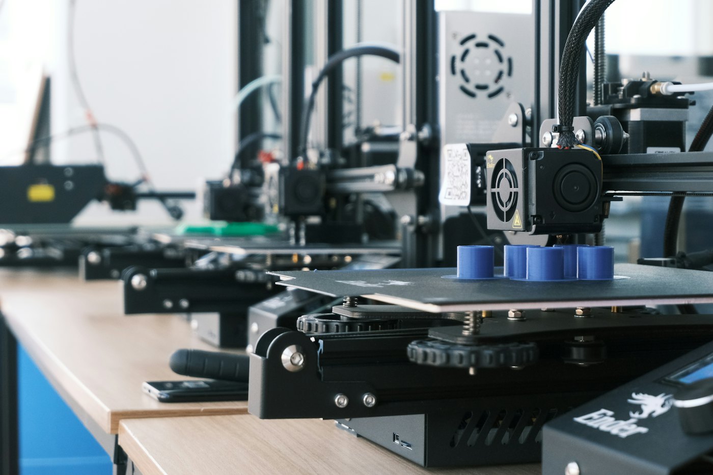 A row of 3D printers making blue plastic parts on a wooden bench
